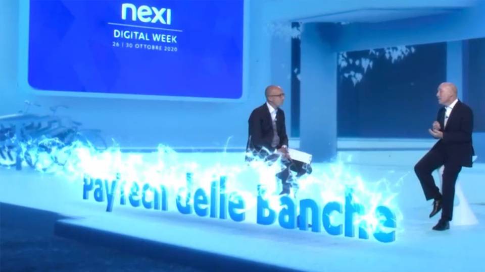 Un momento dell’evento ‘Nexi Digital Week’ con due relatori che discutono, seduti sopra un’animazione digitale che raffigura la scritta ‘PayTech delle Banche’, in uno spazio virtuale dall’atmosfera high-tech.