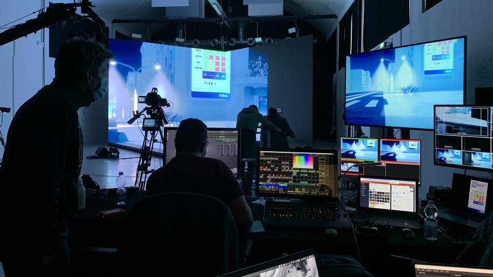 Backstage dell’evento ‘Nexi Digital Week’, con i tecnici impegnati nella gestione delle riprese e delle grafiche virtuali su più monitor di controllo, in uno studio attrezzato con telecamere e schermi.