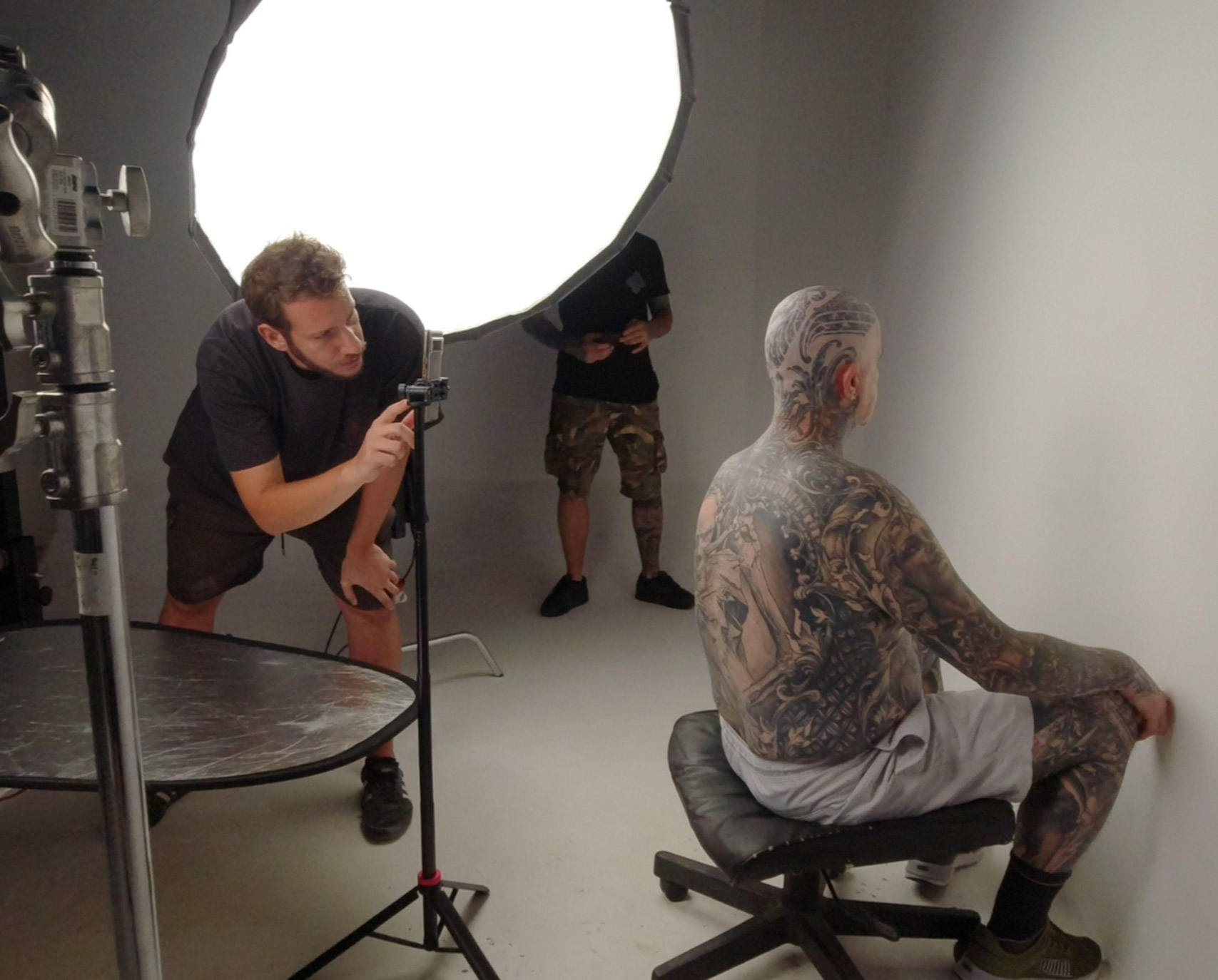 Scatto nel backstage di uno studio fotografico con tecnici al lavoro, mentre riprendono l’uomo tatuato adoperato come modello per l'avatar virtuale seduto su una sedia.